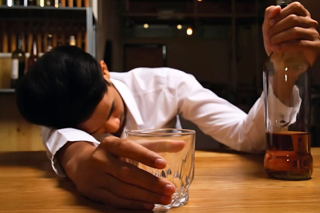 A young man passed out at a bar table with an alcohol bottle in hand, symbolizing alcohol addiction and the need for recovery.